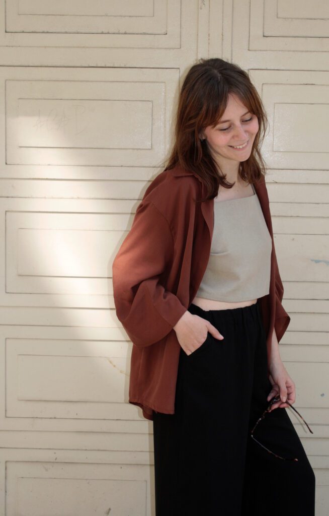 A woman standing in front of a patterned wall wearing a blouse in maroon, a beige linen top and wide black tencel pants. She has one hand in the pocket, with the other one she is holding her sunglasses.