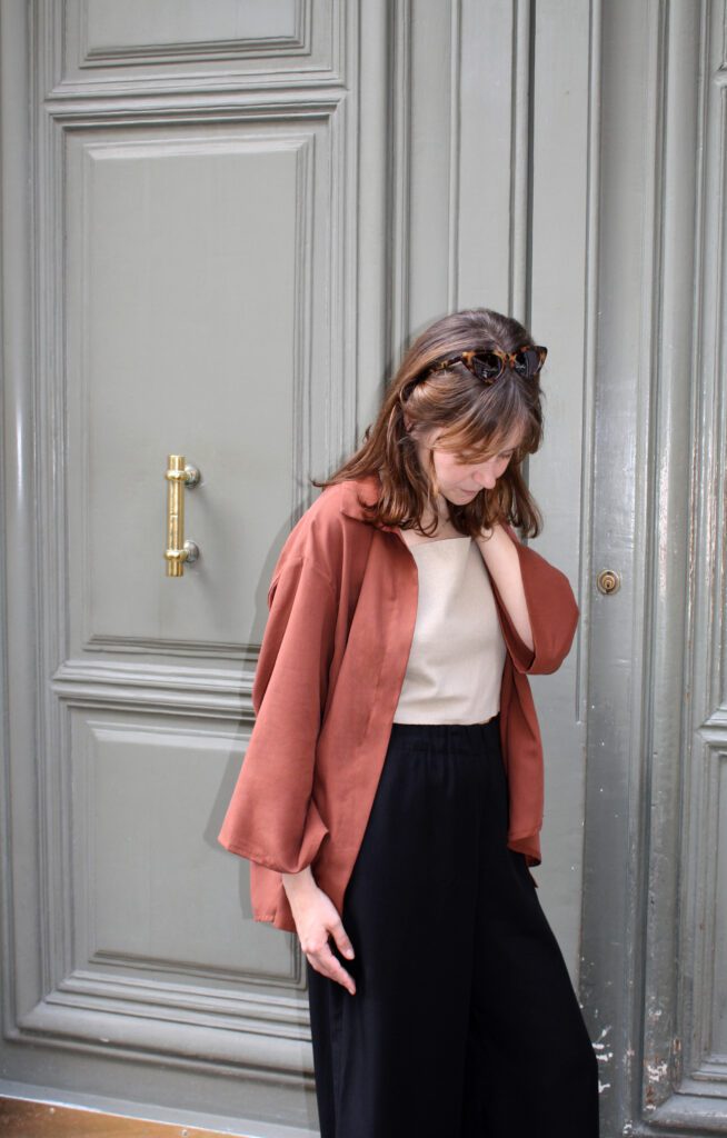 A woman standing in front of a blue-grey door. She is touching her hair and is wearing a maroon-coloured blouse, a linen top and black wide tencel pants.