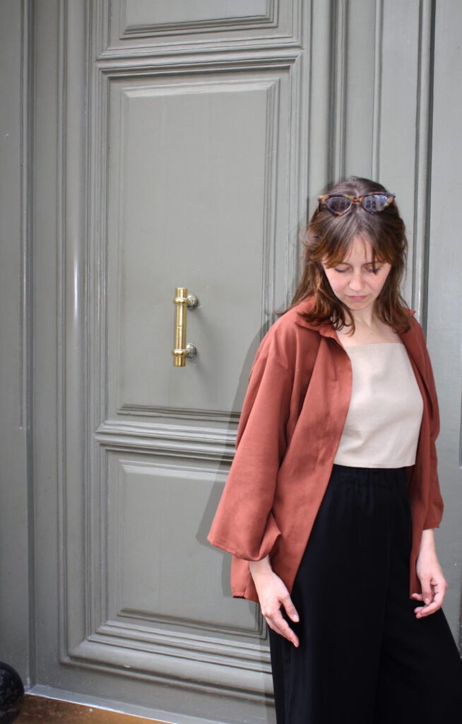 A woman standing in front of a blue-grey door. She is wearing a maroon-coloured blouse, a linen top and black wide tencel pants.