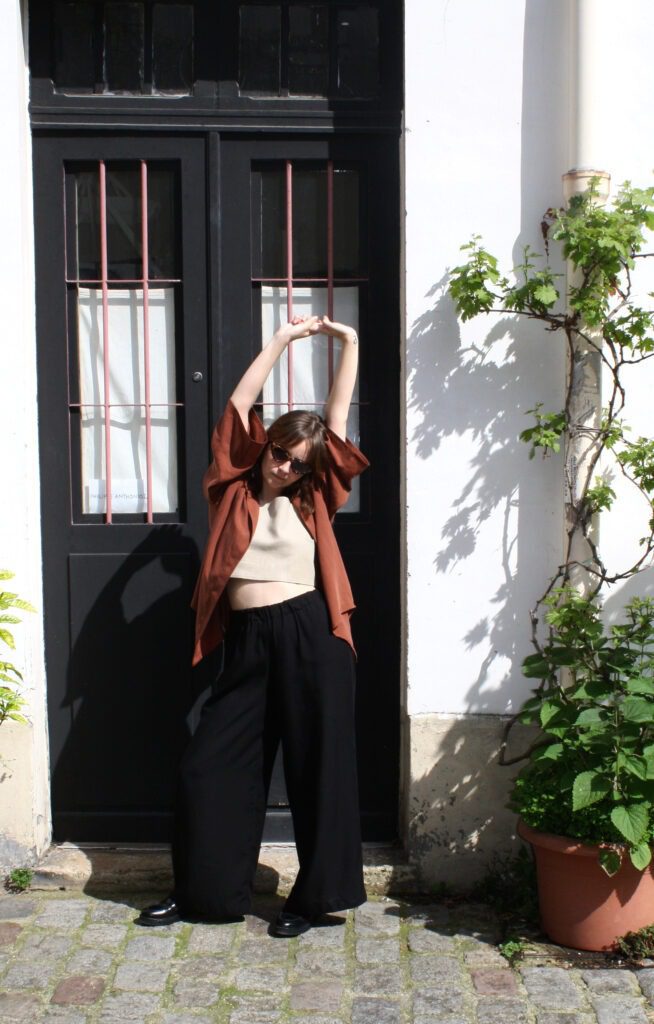 A woman standing in front of a black door next to plants, stretching her arms upwards. She is wearing a maroon-coloured blouse, a linen top and black tencel pants.