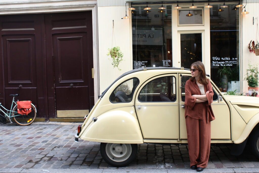 A woman wearing a set of matching maroon-coloured blouse and pants. She is standing in front of an old car with her arms crossed in a beautiful street in Paris.