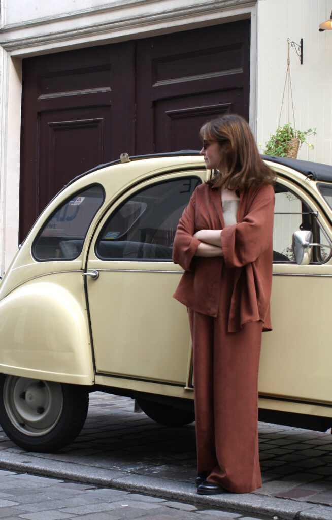 A woman wearing a set of matching maroon-coloured blouse and pants, standing in front of a beige old-timer car in the streets of Paris.